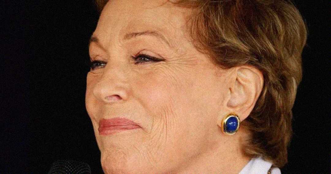 A close-up of an older woman with short light brown hair, wearing blue and gold earrings, smiling gently, and holding a microphone.