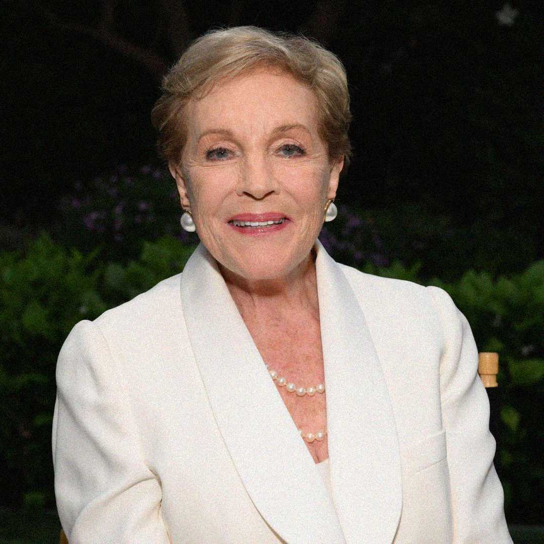 An older woman with short, light hair is smiling. She is wearing a white suit jacket, pearl necklace, and pearl earrings, seated outdoors with greenery in the background.