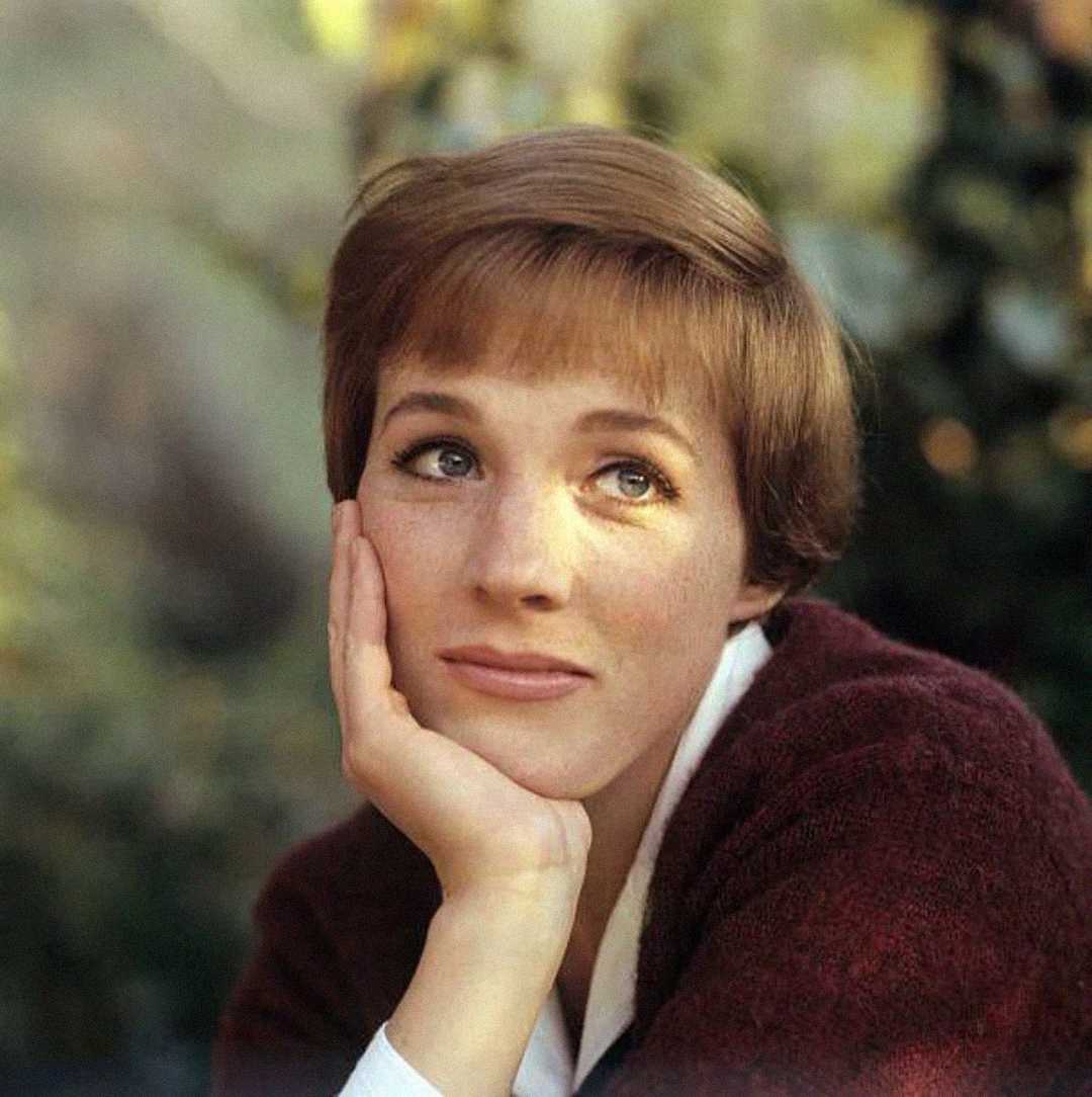 A woman with short brown hair rests her chin on her hand, gazing upwards thoughtfully. She wears a burgundy sweater and sits against a blurred, natural outdoor background.