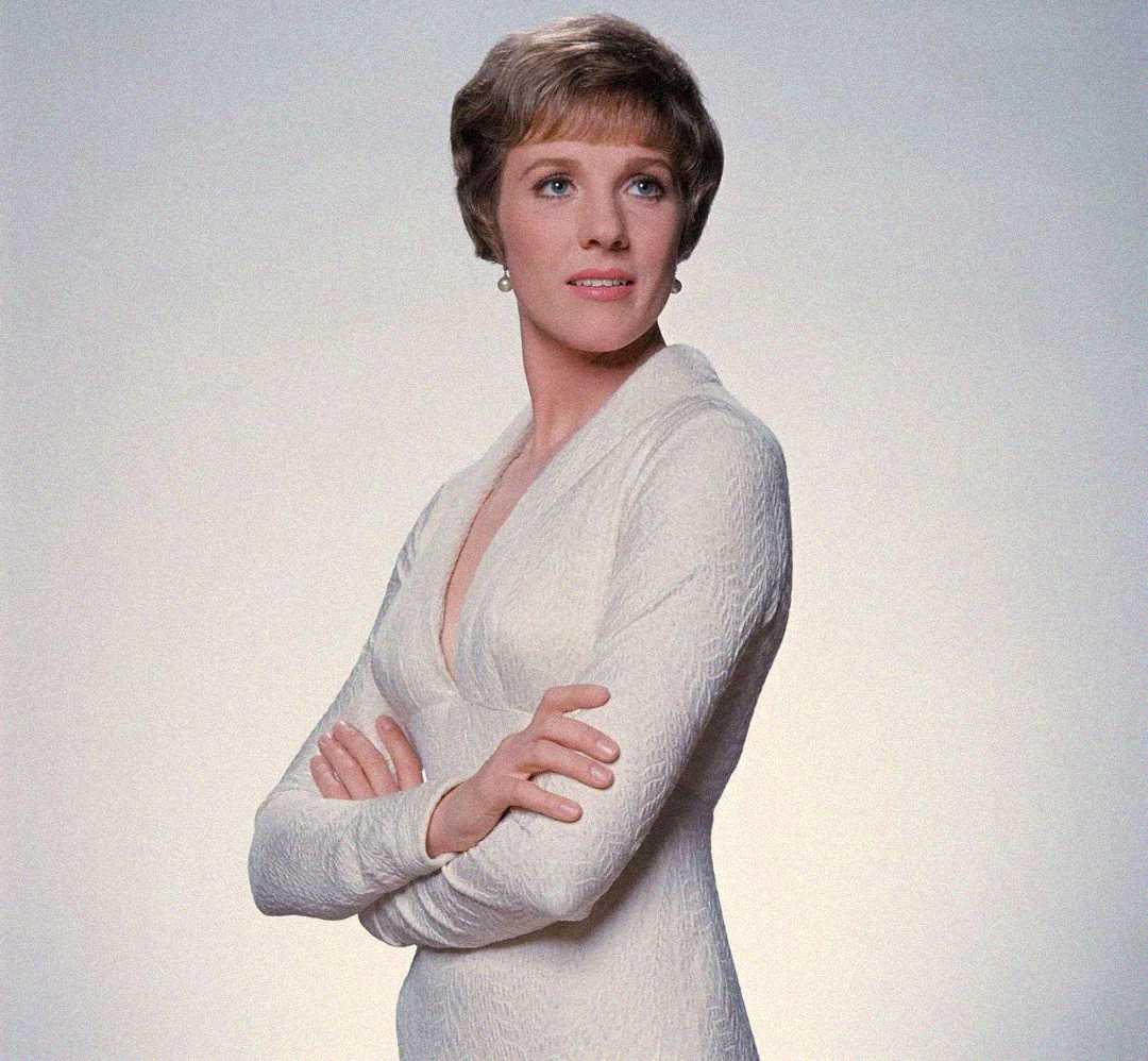 A woman with short light brown hair, wearing a long-sleeved white dress, stands with her arms crossed and looks slightly to the side against a plain light background.