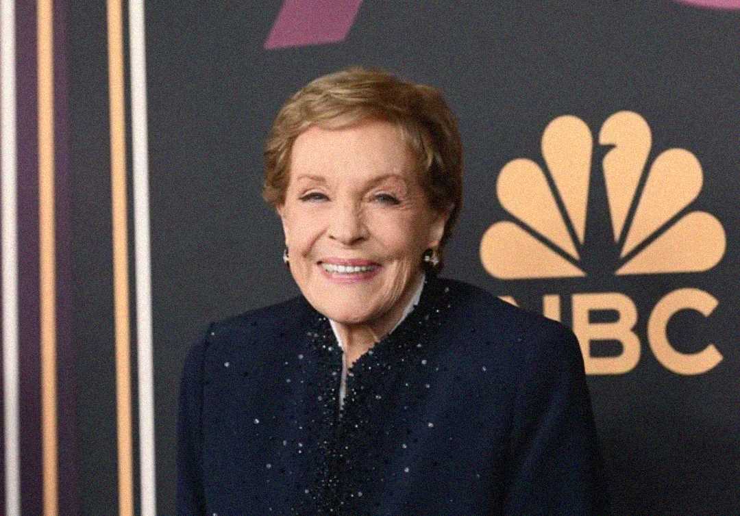 An older woman with short, light brown hair and a navy blue outfit smiles while standing in front of a black backdrop featuring the NBC logo in gold.