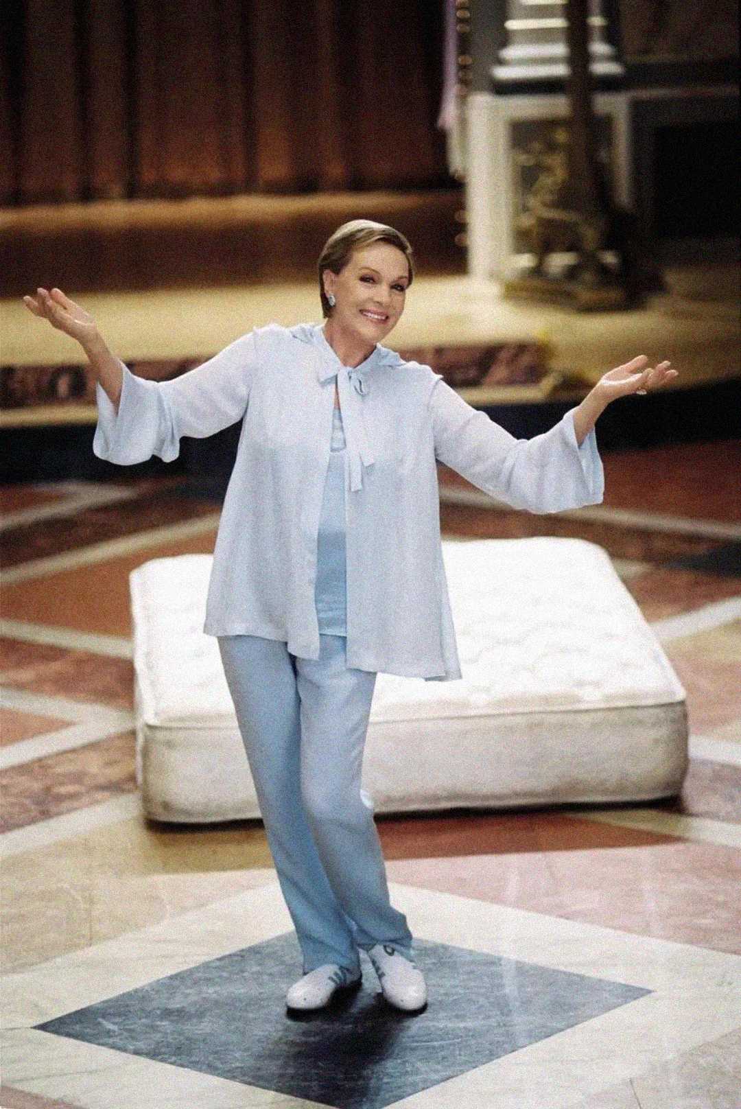 A woman wearing light blue pants and a matching top stands smiling with arms spread wide in front of a mattress on a marble floor, in a large indoor space with ornate decor.
