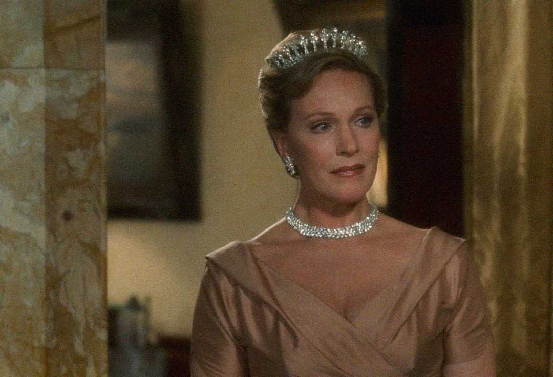 A woman wearing a tiara and diamond necklace stands in an elegant setting, dressed in a champagne-colored gown with cap sleeves and an elegant updo. She appears composed and regal.