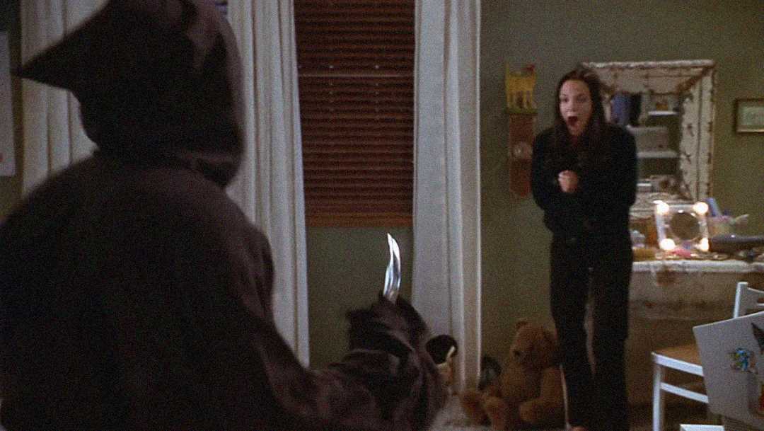 A person dressed in a black hooded costume holds a knife, confronting a terrified woman standing in a bedroom with her hands near her face. Toys and a desk are visible in the background.