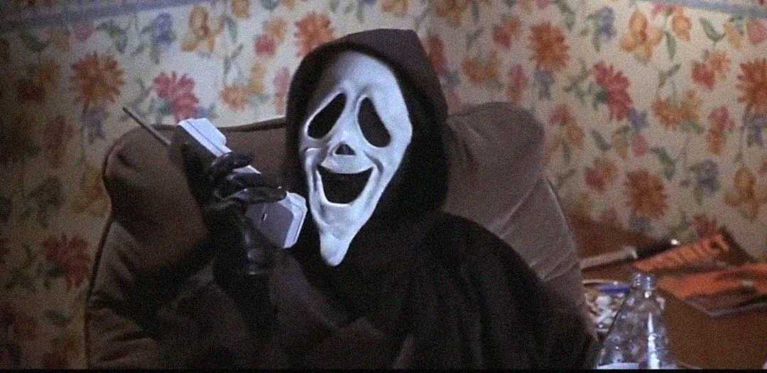 A person wearing a "Ghostface" mask and black hooded robe is sitting indoors, holding a cordless phone to their ear, with floral wallpaper and a cluttered table in the background.