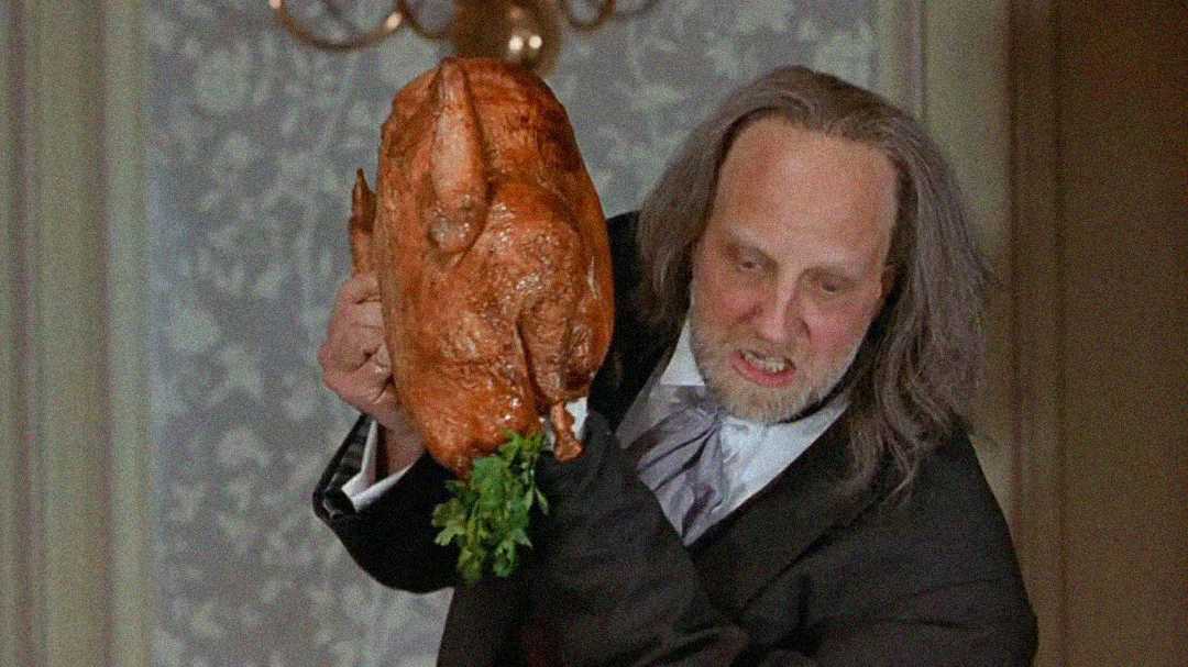A man with long, messy gray hair and a suit angrily raises a large roasted turkey with herbs inside, as if about to throw it, in a room with patterned wallpaper and a chandelier.