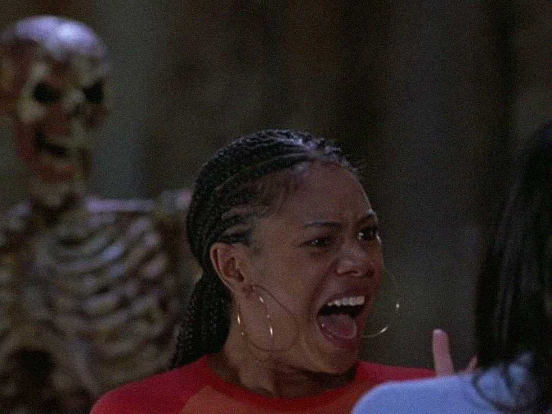 A woman with braided hair and large hoop earrings screams in fear, with her mouth open wide. Behind her, a skeleton stands in the background, partially blurred.