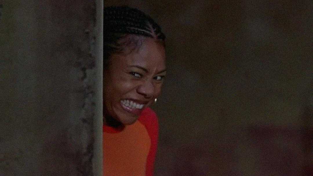 A woman with braided hair and hoop earrings grins mischievously while peeking from behind a wall, wearing an orange top.