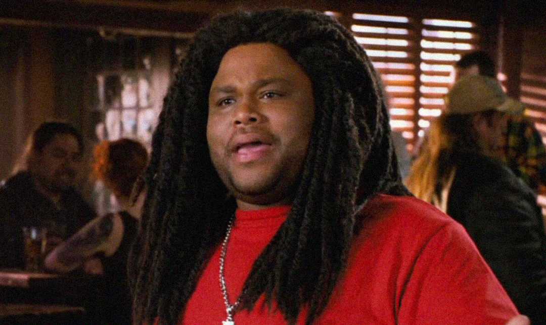 A man with long, dark dreadlocks wearing a red shirt and silver necklace stands indoors, speaking. The background shows other people and wooden blinds letting in some light.