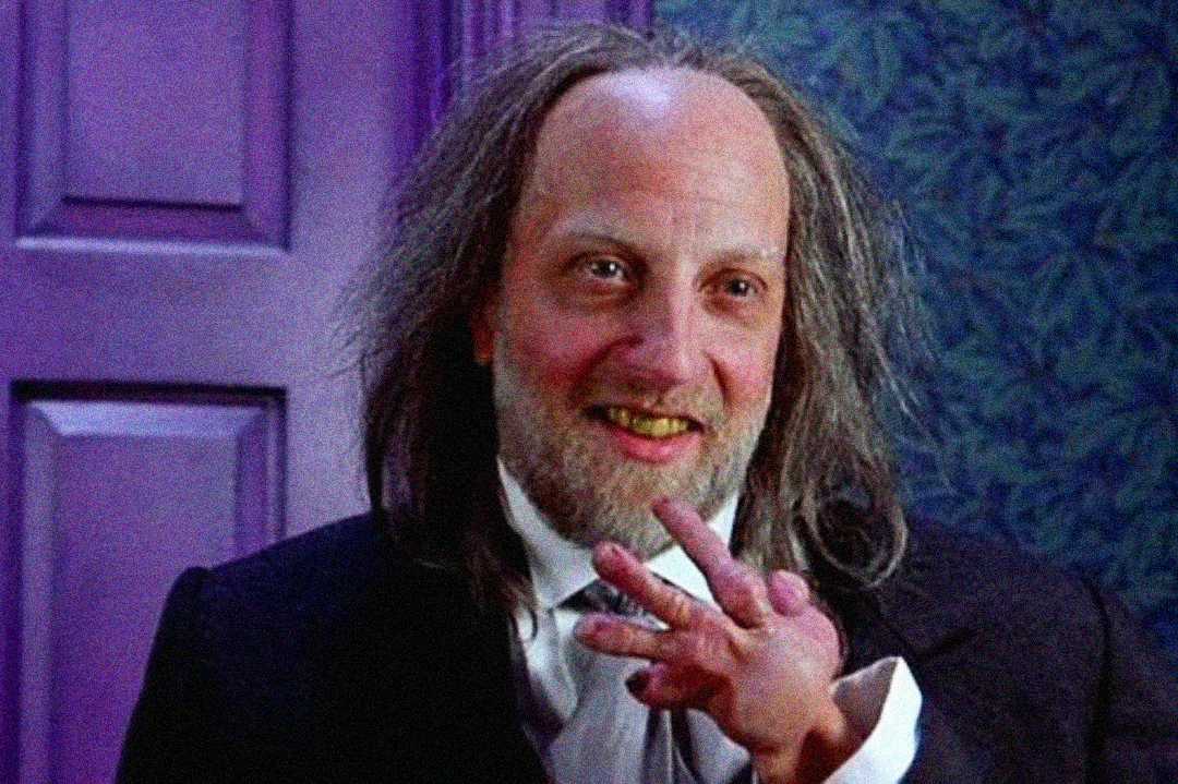 A man with long, messy hair and a receding hairline smiles while holding up a small, deformed hand. He is dressed in a suit and standing indoors against a purple door and patterned wall.