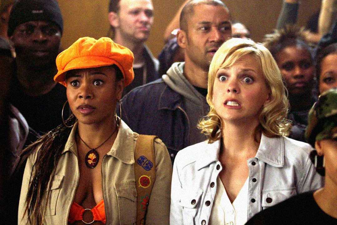 Two women stand close together in a crowd, both looking surprised and concerned. One wears an orange cap and tan jacket, while the other has blonde hair and a white jacket. People are gathered behind them, mostly looking forward.