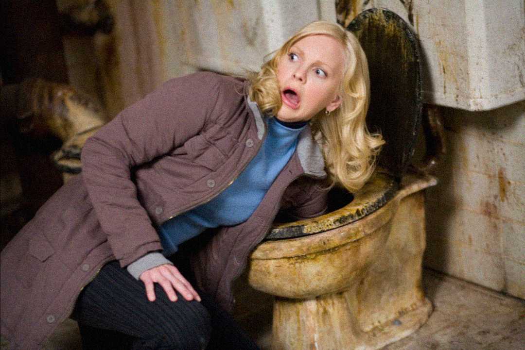 A woman with blonde hair and a shocked expression leans close to a dirty, stained toilet in a grimy bathroom. She is wearing a brown coat and blue top.