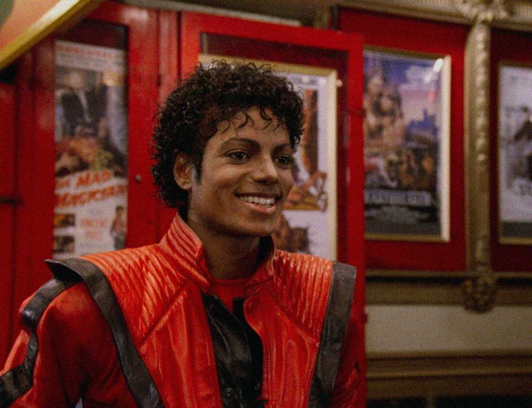 A man with curly hair wearing a red and black leather jacket smiles in front of movie posters in a theater lobby.