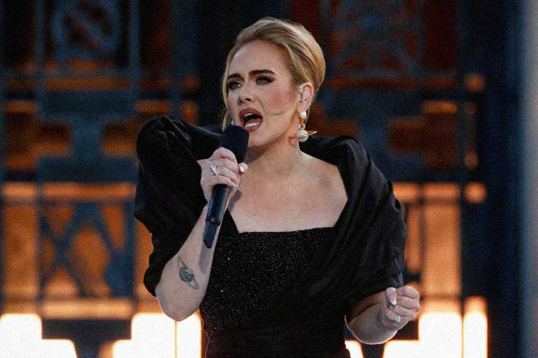 A woman with light hair styled up, wearing a black off-shoulder dress, sings passionately into a microphone on stage with blurred decorative background lighting.