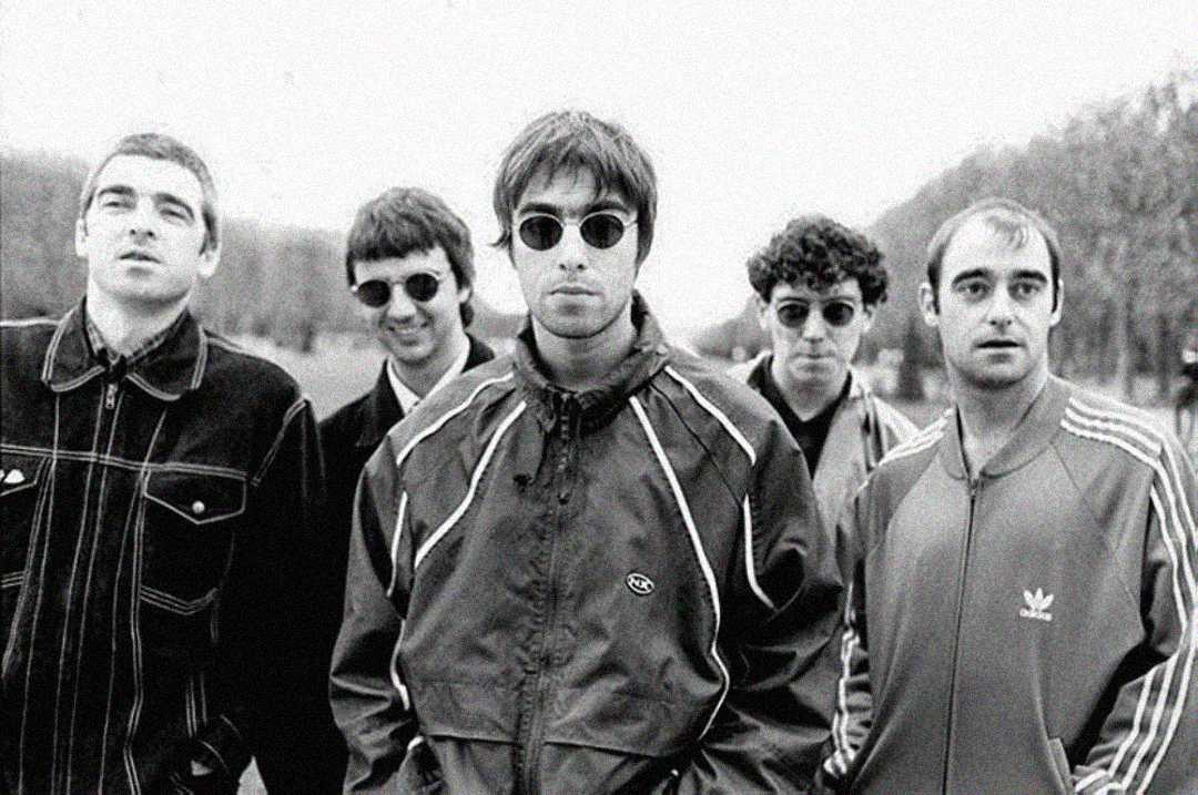 Five men stand outdoors in a black-and-white photo. They wear casual jackets and sunglasses, looking toward the camera with neutral or serious expressions. Trees line the background under an overcast sky.