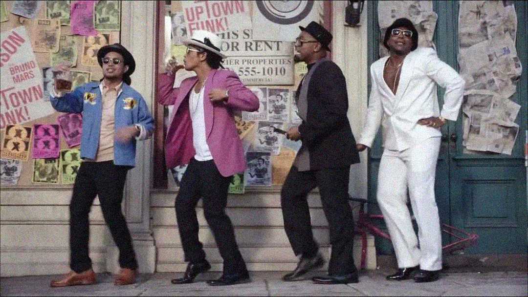 Four men are dancing on a city sidewalk in colorful retro-style outfits, including hats and sunglasses, in front of a wall covered with posters and notices. One man wears a white suit while the others wear casual jackets.