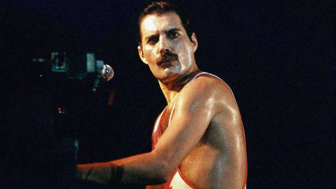 A person with short dark hair and a mustache, wearing a sleeveless top, is sweating and playing keyboard on stage, looking toward the camera under bright stage lights.