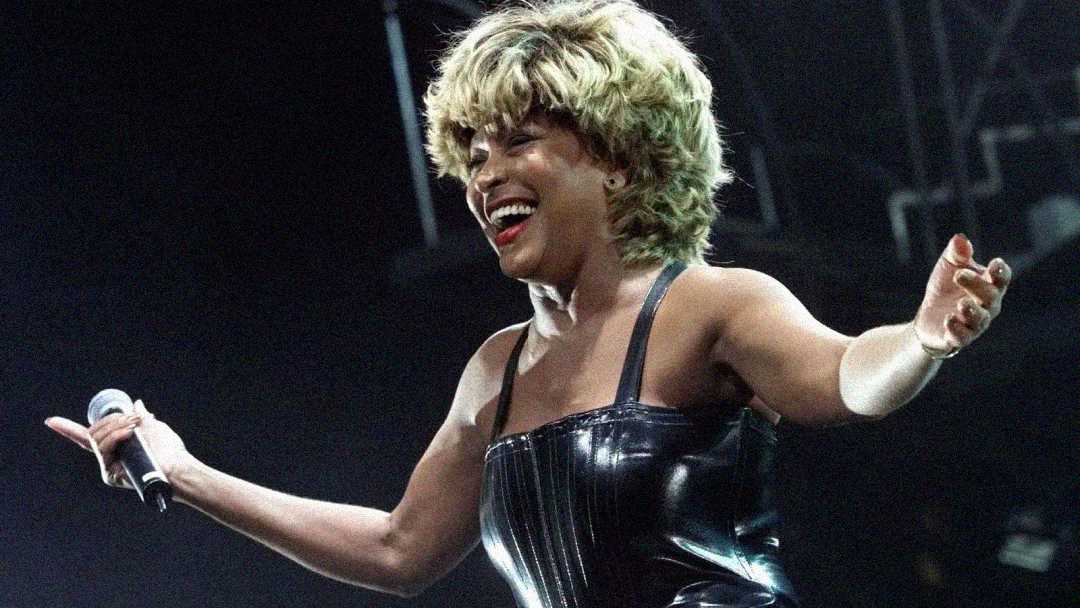 A woman with short, blonde hair and a black, sleeveless leather outfit smiles and holds a microphone while performing on stage under bright lights.