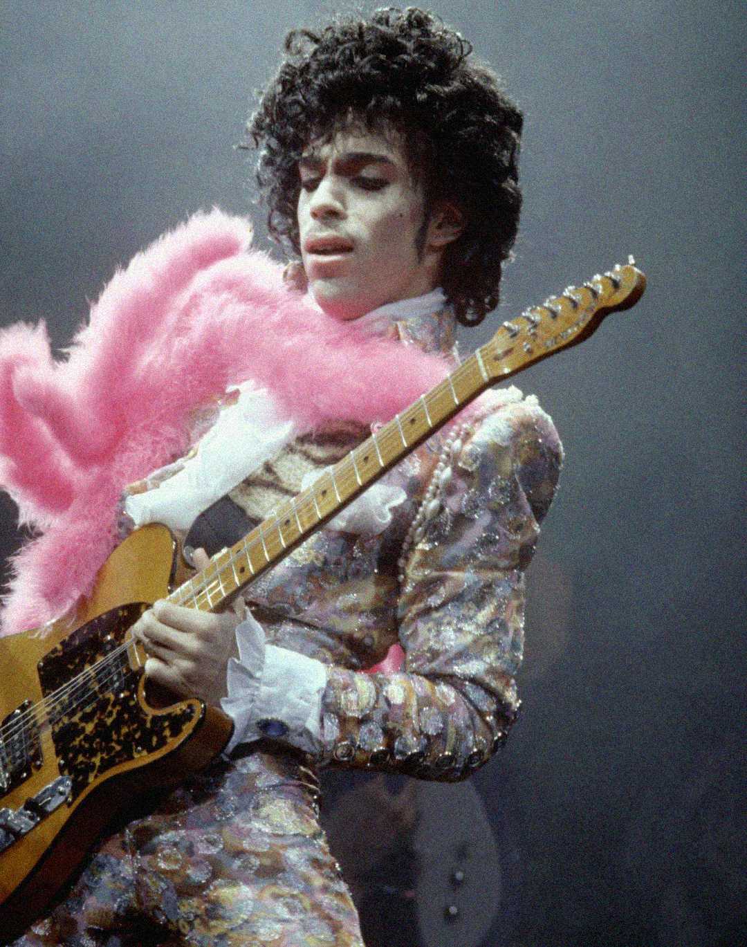 A musician wearing a glittery, patterned suit and a pink feather boa plays an electric guitar passionately on stage, surrounded by dramatic stage lighting.