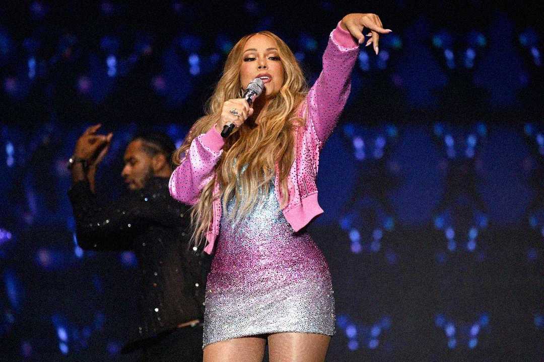 A woman wearing a sparkling pink and silver dress with a pink jacket performs on stage, holding a microphone and pointing toward the audience. Another performer is visible in the background against a dark, lit-up backdrop.