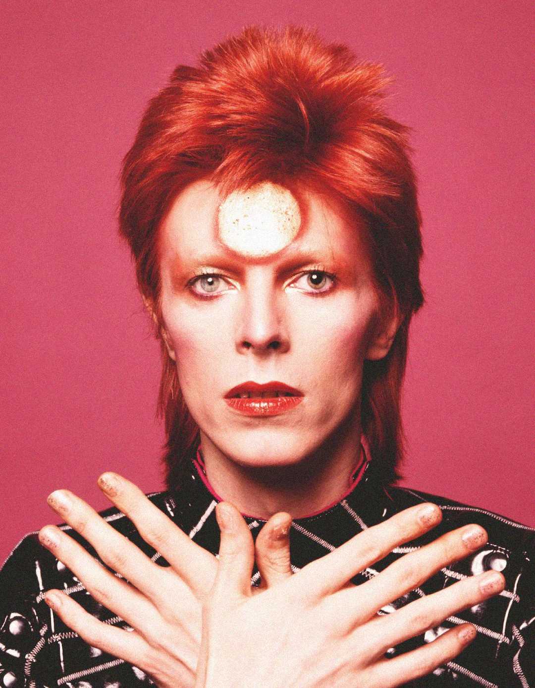 Person with pale skin and bright red hair styled upward, wearing bold makeup and a red or pinkish lipstick. They have a circular metallic object on their forehead and are posing with crossed hands in front of a pink background.
