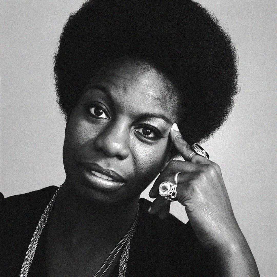 Black and white portrait of a woman with an afro hairstyle, wearing rings and a necklace, resting her head on her hand with a thoughtful expression.
