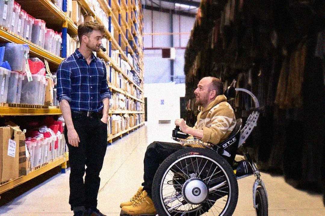A man in a wheelchair talks with a standing man in a plaid shirt inside a warehouse lined with shelves full of labeled boxes and containers.