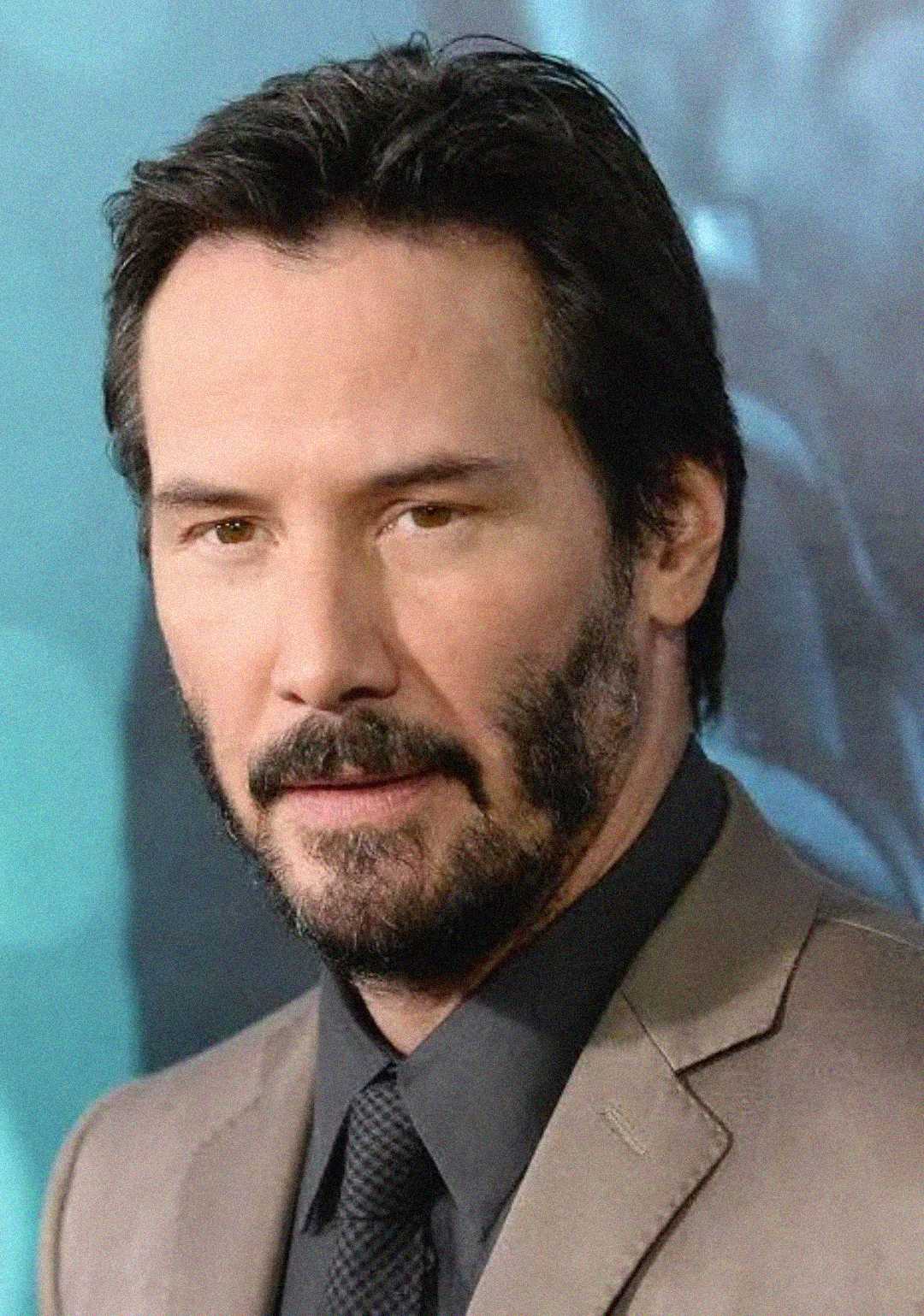 A man with dark hair and a beard, wearing a gray suit jacket, black shirt, and black tie, stands in front of a blurred blue-toned background.