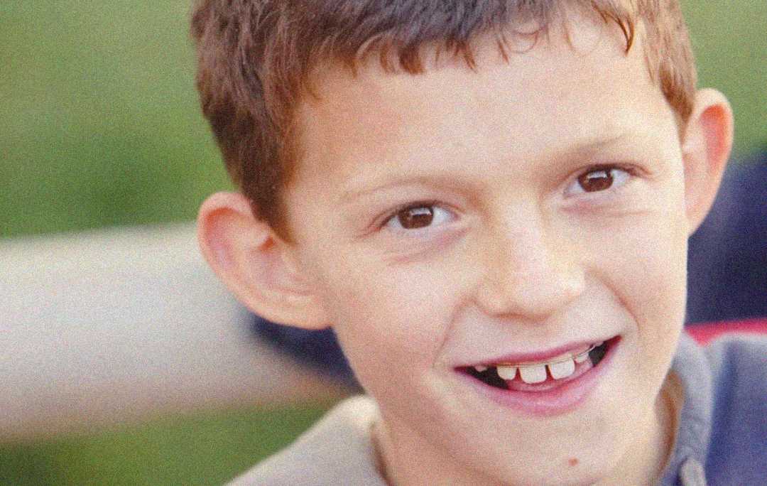 A young boy with short brown hair smiles at the camera, showing a gap between his front teeth. The background is blurred with green tones.