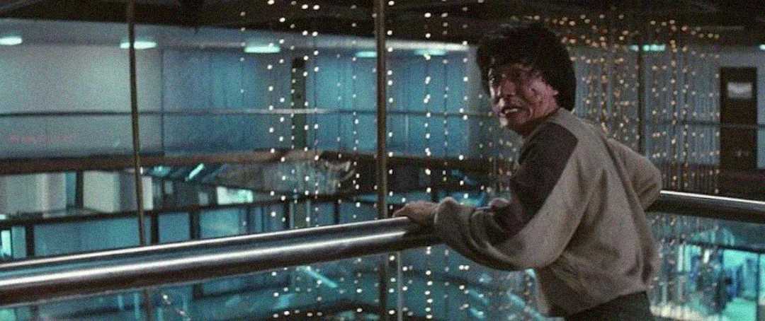 A man with disheveled hair and a bruised face grips a railing inside a building decorated with strings of small white lights, looking intensely to the side.