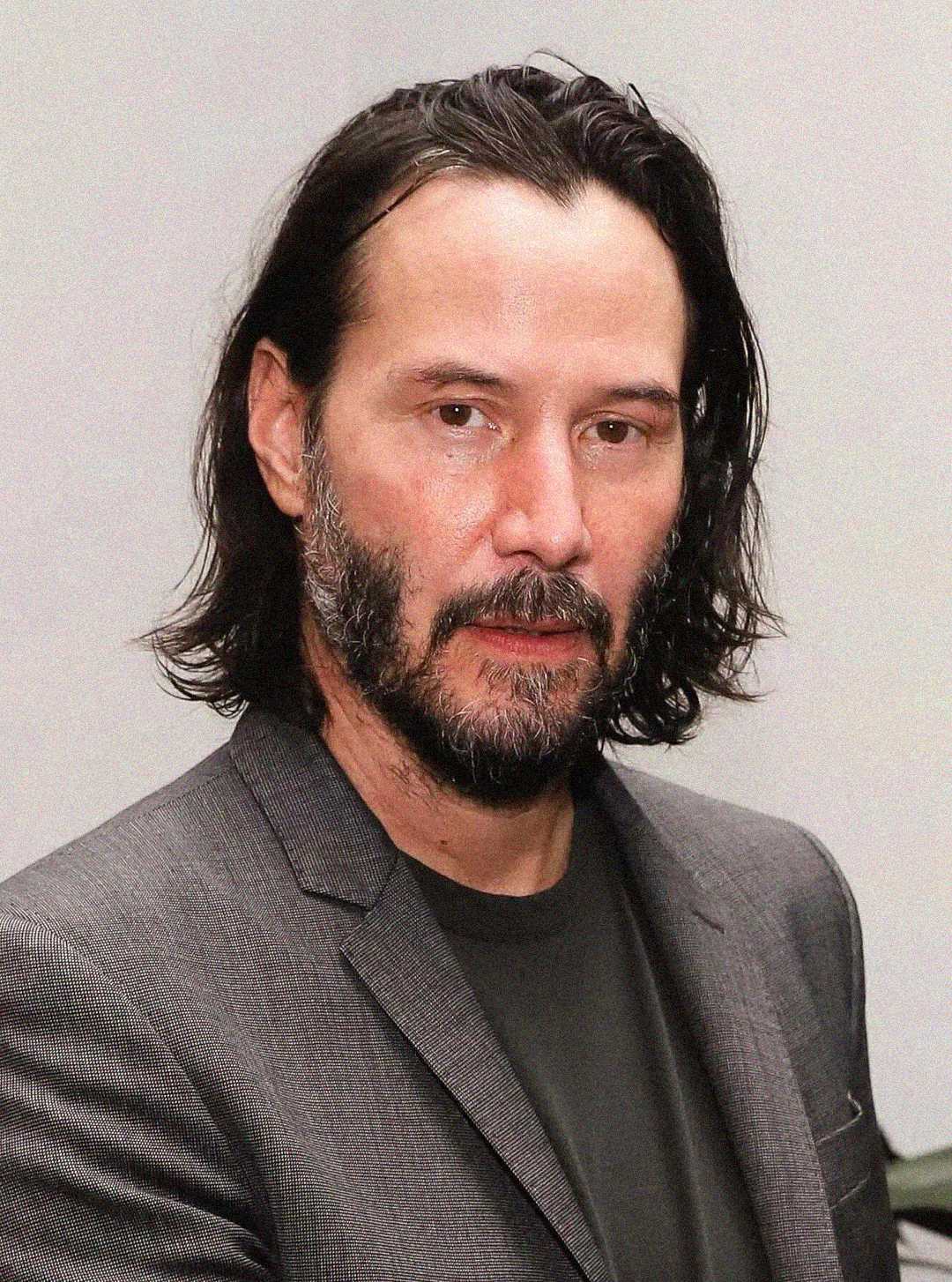 A man with shoulder-length dark hair, a beard, and mustache, wearing a gray blazer over a dark shirt, poses against a plain light background.