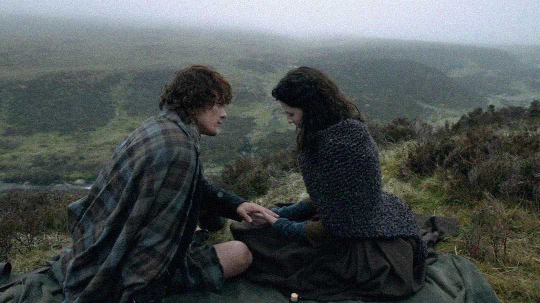A man and woman sit closely on a blanket outdoors in a misty, hilly landscape. They face each other, holding hands, dressed in old-fashioned, warm clothing. The scene is intimate and calm, surrounded by nature.