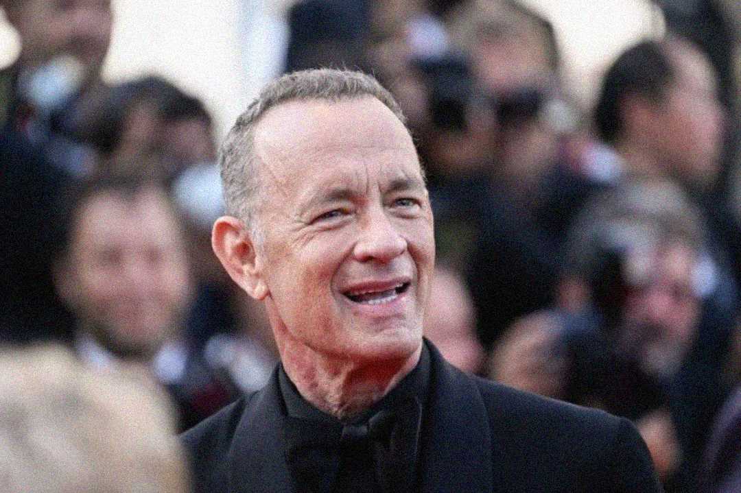 A man in a black tuxedo smiles while standing among a crowd at a formal event, with people and cameras visible in the blurred background.
