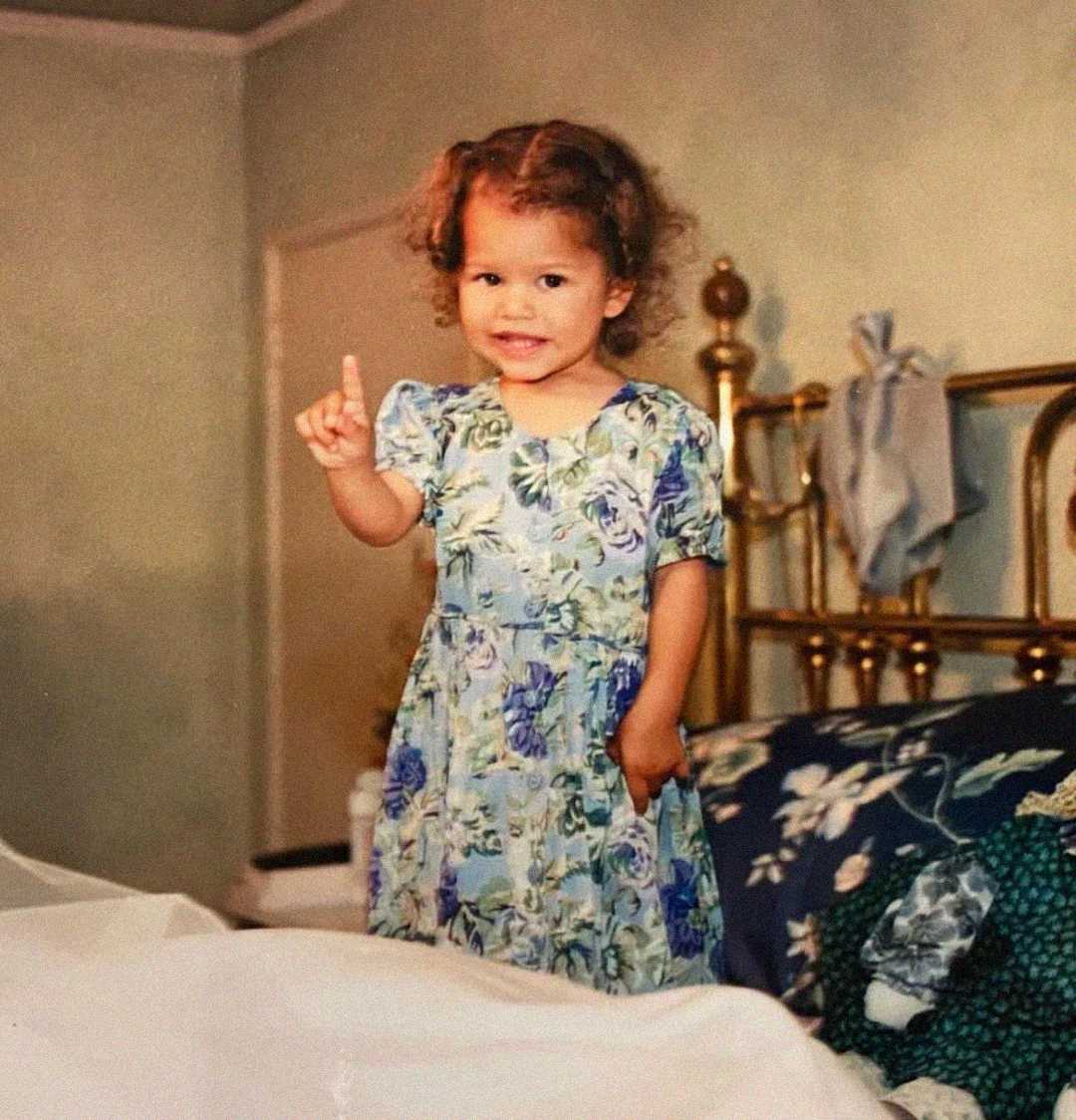 A young child with curly hair, wearing a floral dress, stands on a bed with one finger raised and smiles at the camera. The background shows a brass bed frame and light-colored walls.