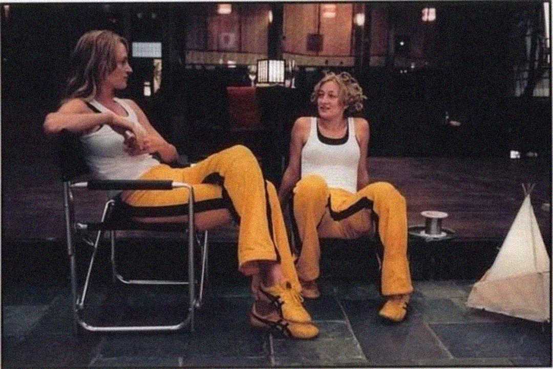 Two women in matching outfits—yellow pants with black stripes and white tank tops—sit and talk in a dimly lit room. One is seated in a chair, the other on the floor, both relaxed and smiling.