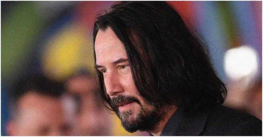 A man with long dark hair and a beard, wearing a dark-colored jacket, stands in front of a blurred, colorful background.