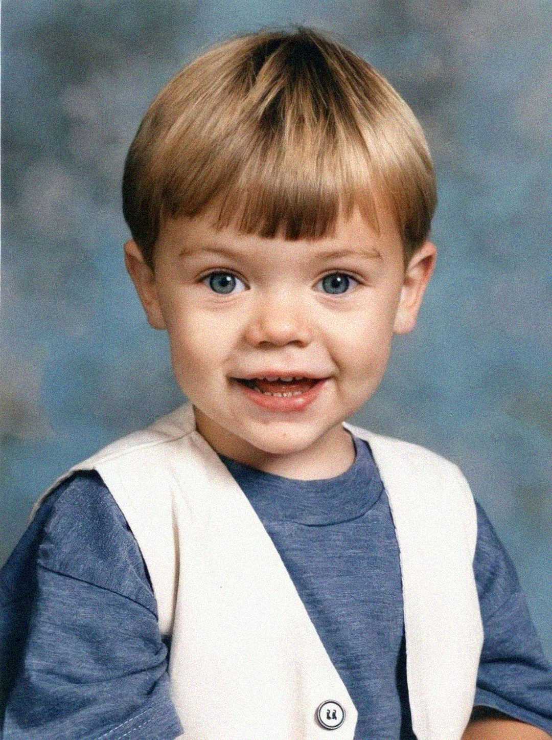 A young child with short blonde hair and blue eyes smiles at the camera. The child is wearing a blue shirt and a light-colored vest, posed in front of a soft, blue-gray studio background.
