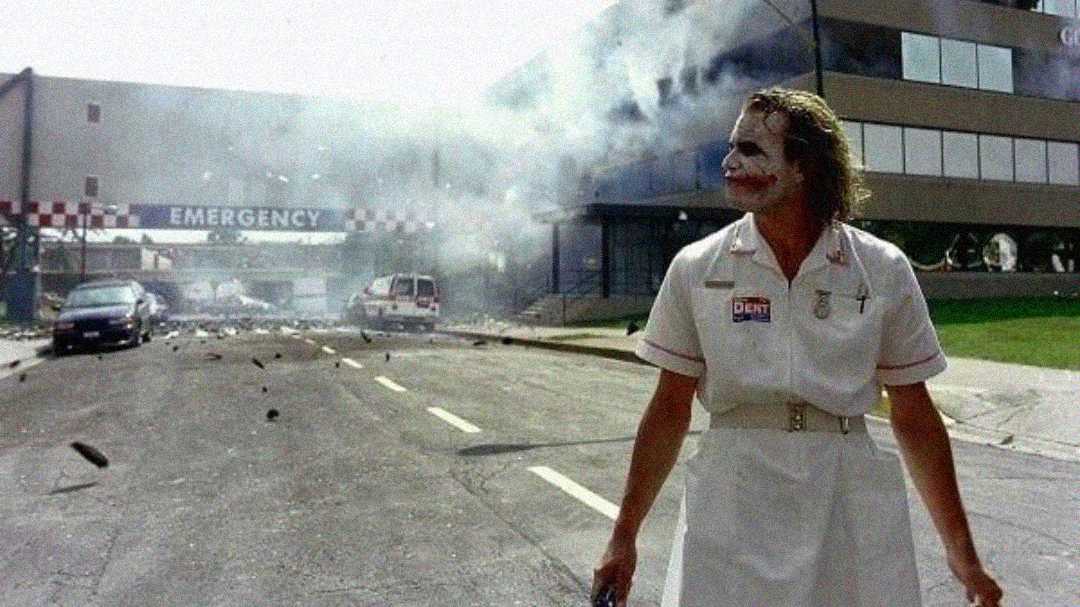 A person in a nurse costume with clown makeup stands on a street in front of a smoking, partially destroyed hospital building, with debris scattered and an "EMERGENCY" sign visible in the background.