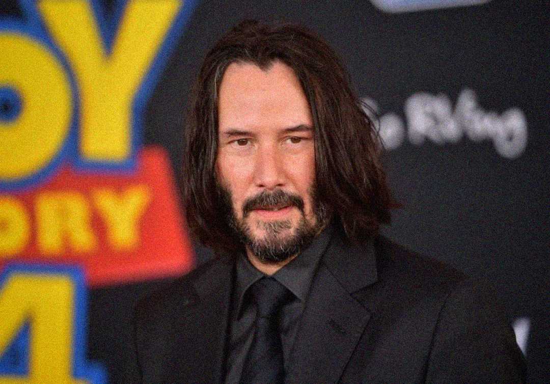 A man with shoulder-length dark hair and a beard, dressed in a black suit, stands in front of a Toy Story 4 movie poster backdrop.