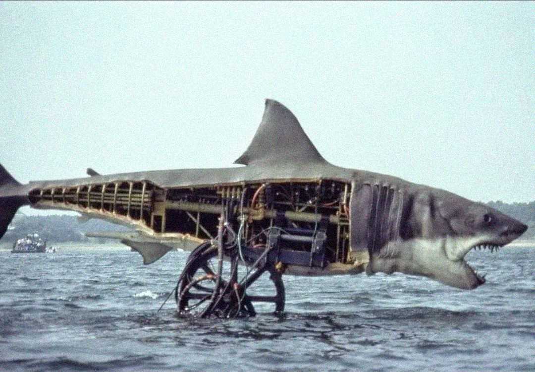 A large mechanical shark with exposed machinery and wires is partially submerged in water, resembling a realistic shark but with its interior visible. The background shows a distant shoreline and a boat.