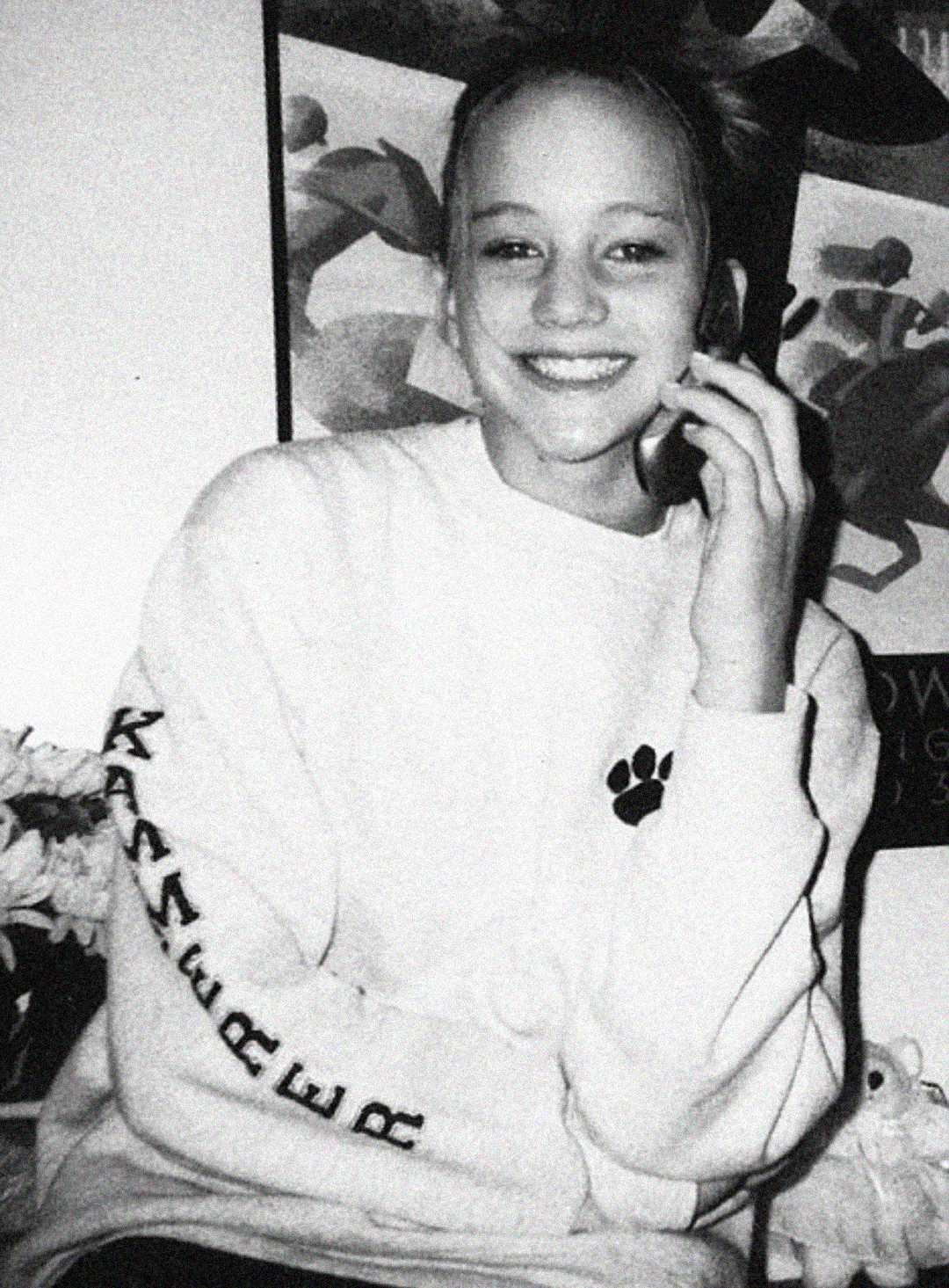 Black and white photo of a smiling young person with hair pulled back, wearing a light sweatshirt with a paw print and text on the sleeve, sitting indoors with one hand touching their face. Poster art is visible in the background.