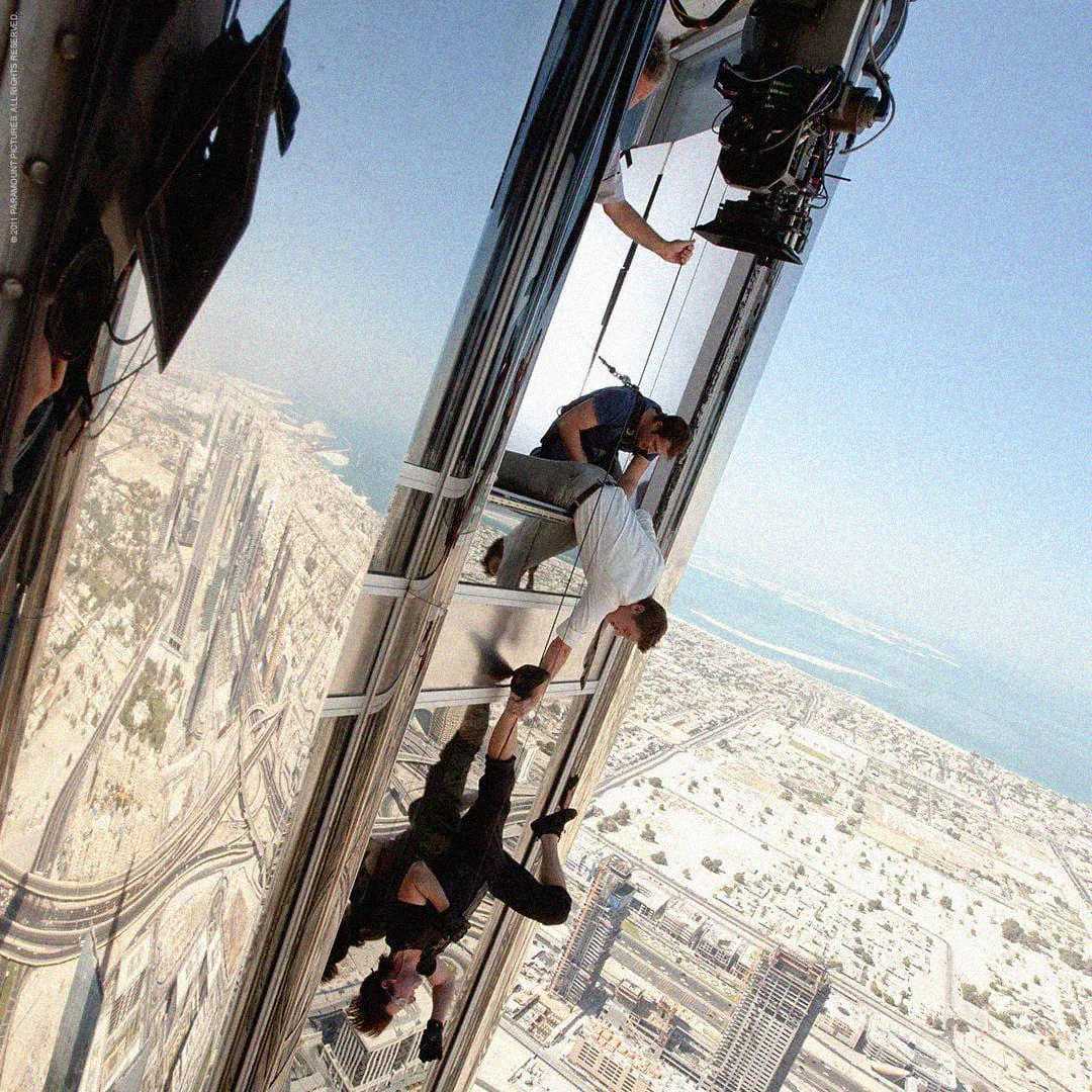 Two people are suspended on the side of a tall glass skyscraper, with a cityscape below. Crew members and camera equipment indicate a film set high above the ground.