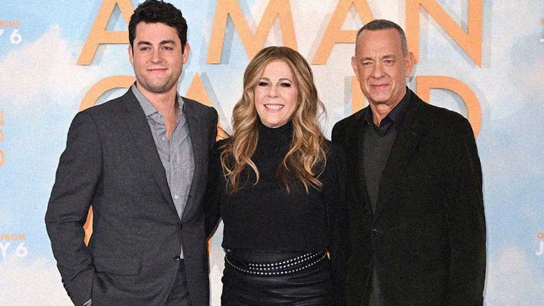 Three adults stand together smiling at a movie premiere, posing in front of a blue and orange backdrop with large text. The man on the left wears a gray suit, the woman in the middle wears black, and the man on the right wears a black coat.