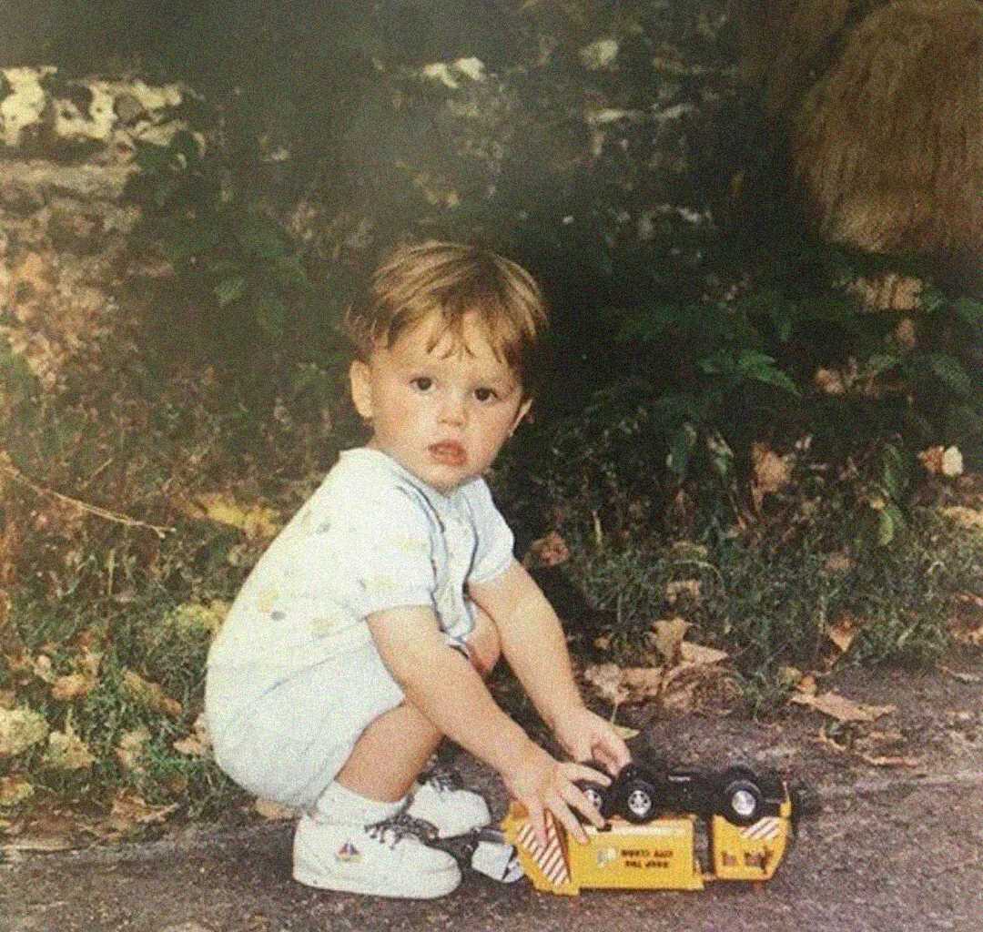 A young child with light brown hair squats outdoors on a path, wearing a light-colored outfit and white sneakers, playing with a yellow toy truck surrounded by plants and fallen leaves.