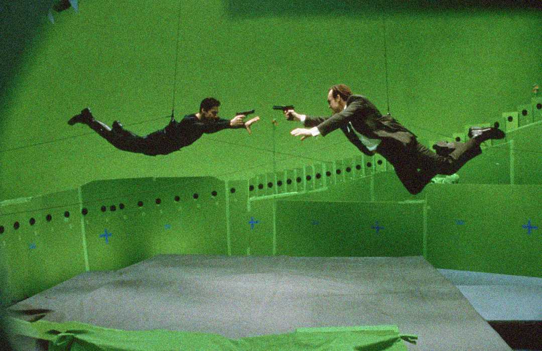 Two actors in action poses, suspended by wires in front of a green screen, point guns at each other in midair on a film set. The background and floor are covered in green panels for visual effects.