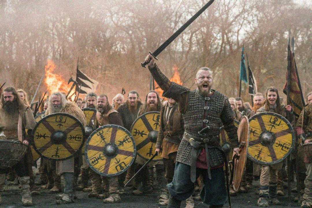 A fierce warrior in armor stands at the front of a group, raising his sword and shouting. Behind him, a crowd of warriors with shields and weapons march forward, with flames and trees in the background.