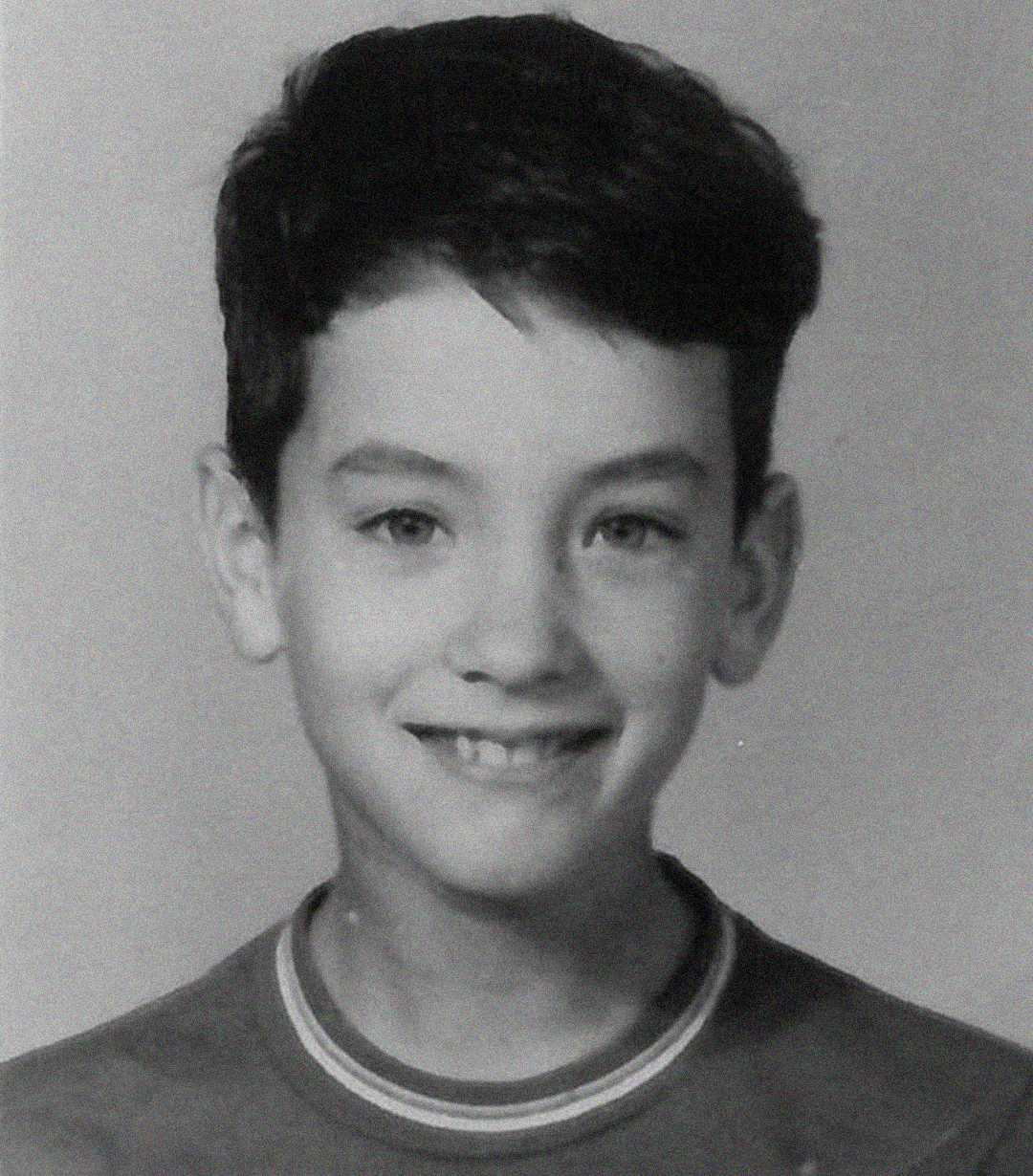 Black and white portrait of a young boy with short dark hair, wearing a crew-neck shirt, smiling at the camera against a plain background.