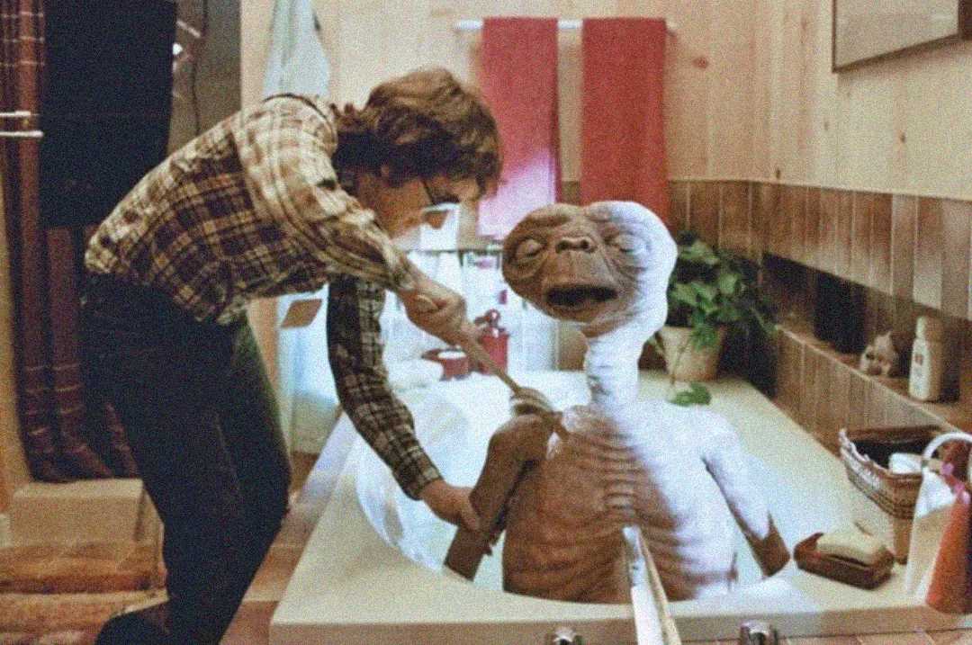 A person in a plaid shirt helps an alien creature, resembling E.T., sit up in a bathtub in a retro-looking bathroom with brown tiles and red towels.