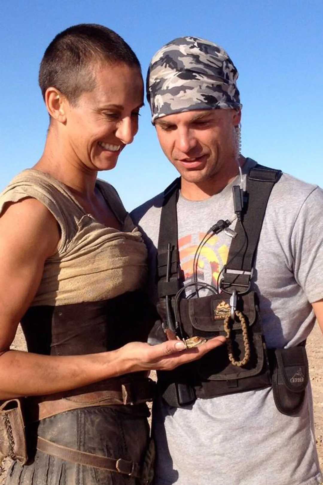 Two people stand close together outdoors under a clear blue sky, smiling and looking at something small resting in one person’s hand. One wears a printed bandana and gear vest; the other has short hair and a sleeveless outfit.