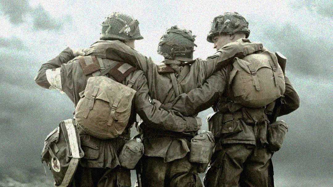 Three soldiers in military gear stand arm-in-arm with their backs to the camera, supporting each other against a cloudy sky. They wear helmets, backpacks, and uniforms, showing camaraderie and solidarity.